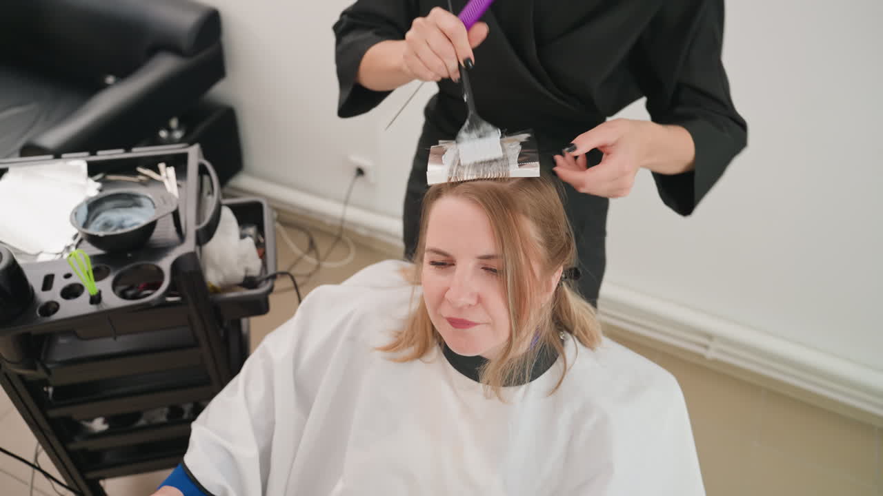 Primo piano di una donna in un salone che fa i colpi di sole, la colorista applica la pellicola e la tinta con un pennello, la cliente indossa una mantellina bianca ed è seduta su una sedia moderna vicino alla macchina per il trattamento, luce naturale soffusa, espressione calma