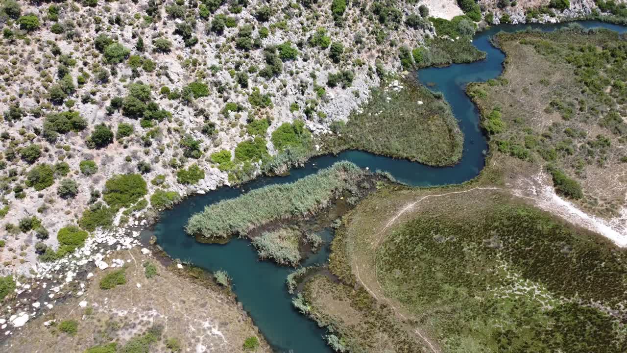 Aerial drone shot showing meandering stream winding through rocky and marshy terrain near Koycegiz Wetlands in Mugla with green patches, textured landscape, calm water bends, and bright daylight