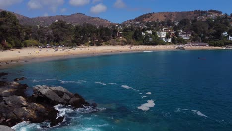 Summer-flight-over-rocks,-waves-and-beautiful-beach-with-mountains-in-the-background