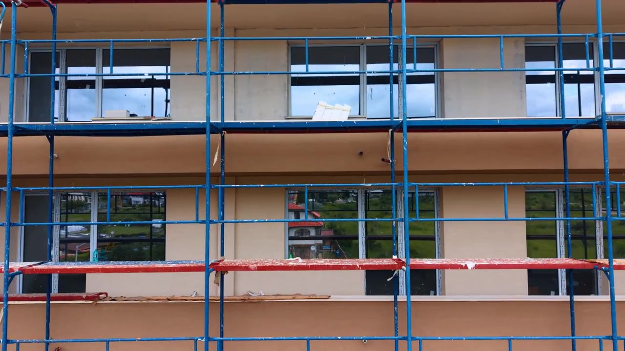 Byggestillads på facade af boligbygning med vinduer. Frontal udsigt over blåt og rødt metalstillads, der dækker en beige bygningsfacade med installerede glasvinduer.