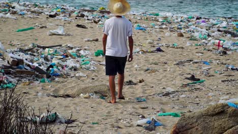 un joven camina por una playa contaminada por basura plástica mirando una instalación industrial