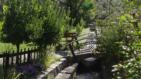 decorative metal bridge over narrow stream surrounded by lush greenery and a wooden bench in a garden setting, montenegro