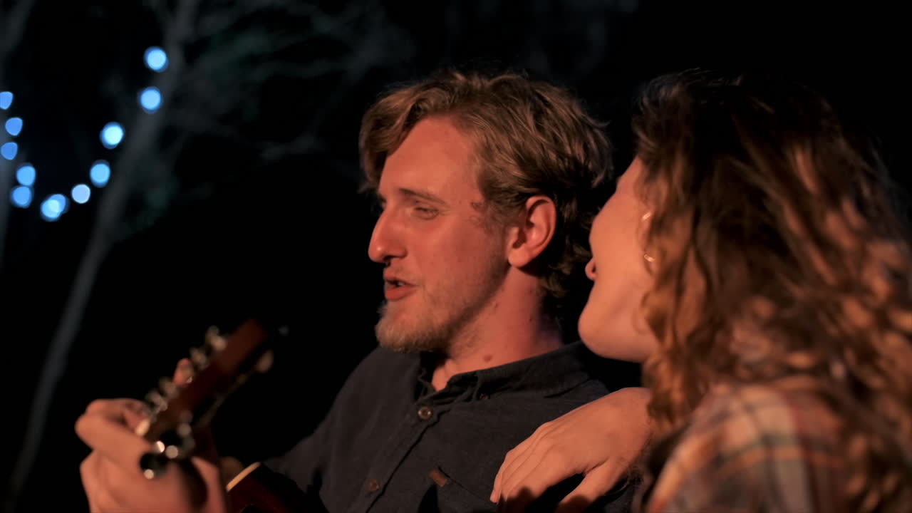 A group of happy young friends near a campfire at glamping, night. Guy playing guitar with woman nearby. Slow motion