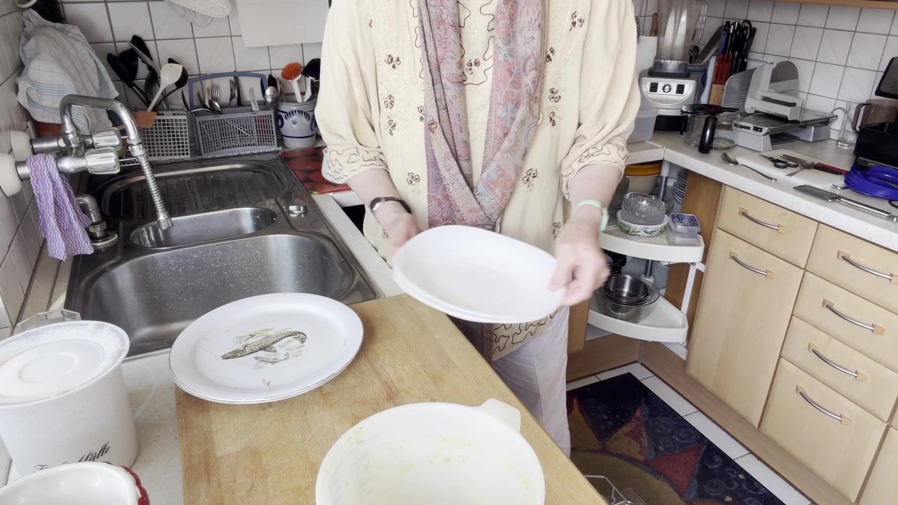 A static kitchen scene where a homemaker steadily transfers decorative fish-patterned plates from a wooden board beside the sink into an open dishwasher in a modern home.