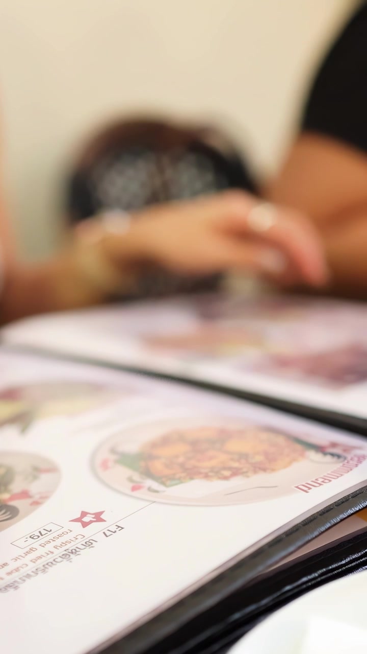 Two people sit together in a well-lit restaurant, flipping through a colorful menu of Northern Thai dishes, discussing meal choices. Shallow depth of field highlights menu details