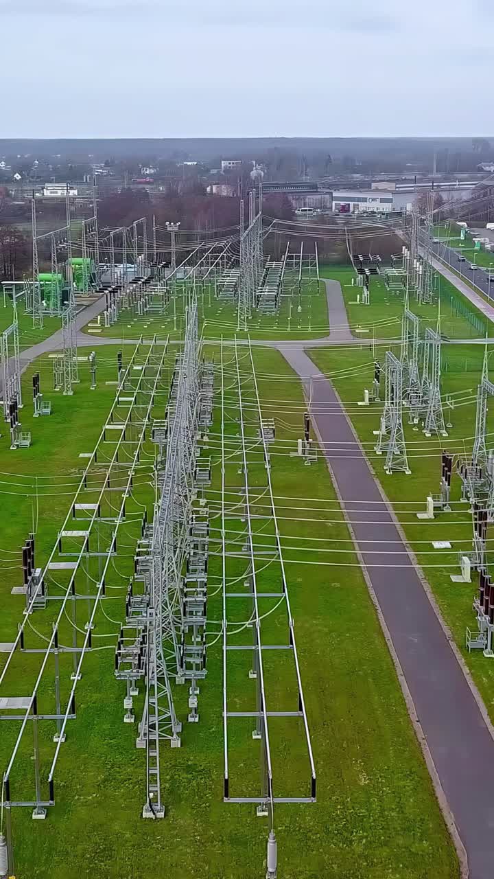 A cinematic parallax drone shot reveals the complex infrastructure of a large electrical substation in Latvia, showing high-voltage power lines, transformers, and pylons that form the energy grid.