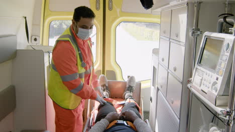Sanitäter-Mit-Gesichtsmaske-Berühren-Das-Knie-Eines-Patienten,-Der-Auf-Der-Trage-In-Einem-Krankenwagen-Liegt