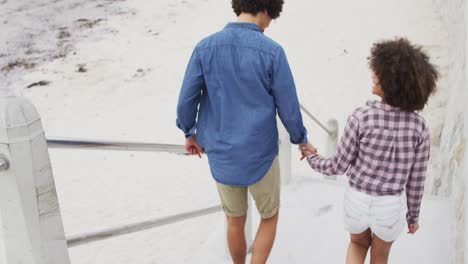 Rear-view-of-african-american-couple-holding-hands-climbing-down-the-stairs-near-the-beach