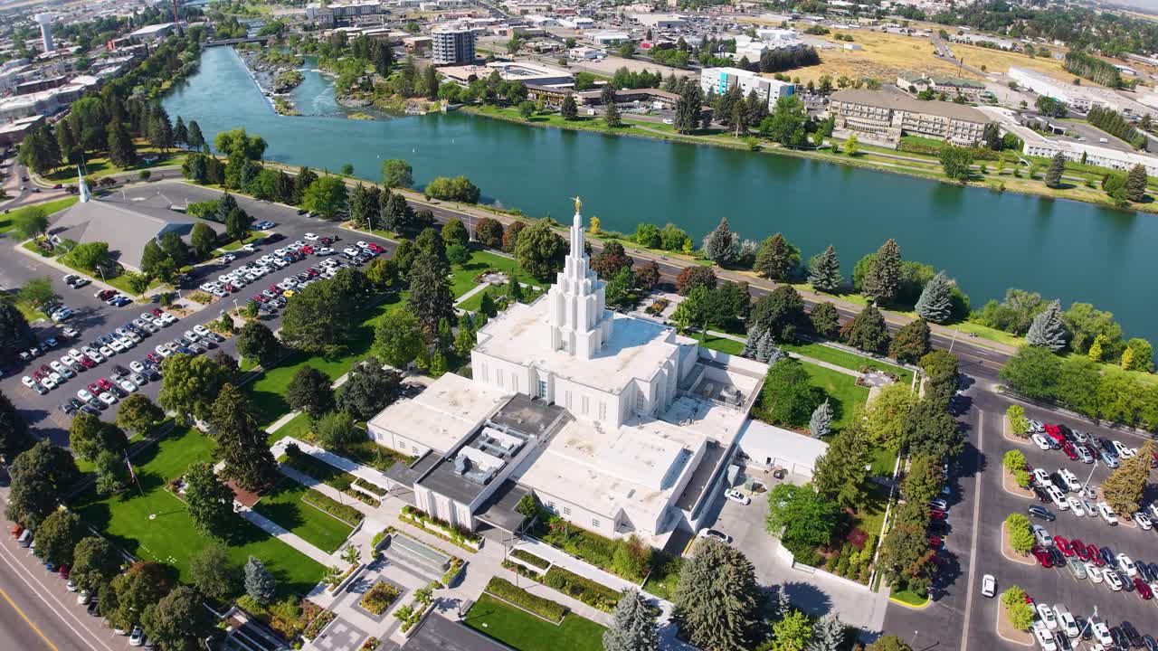 Filmato aereo di un tempio bianco con una guglia alta circondato da giardini verdi, parcheggio e strade accanto al fiume Snake a Idaho Falls