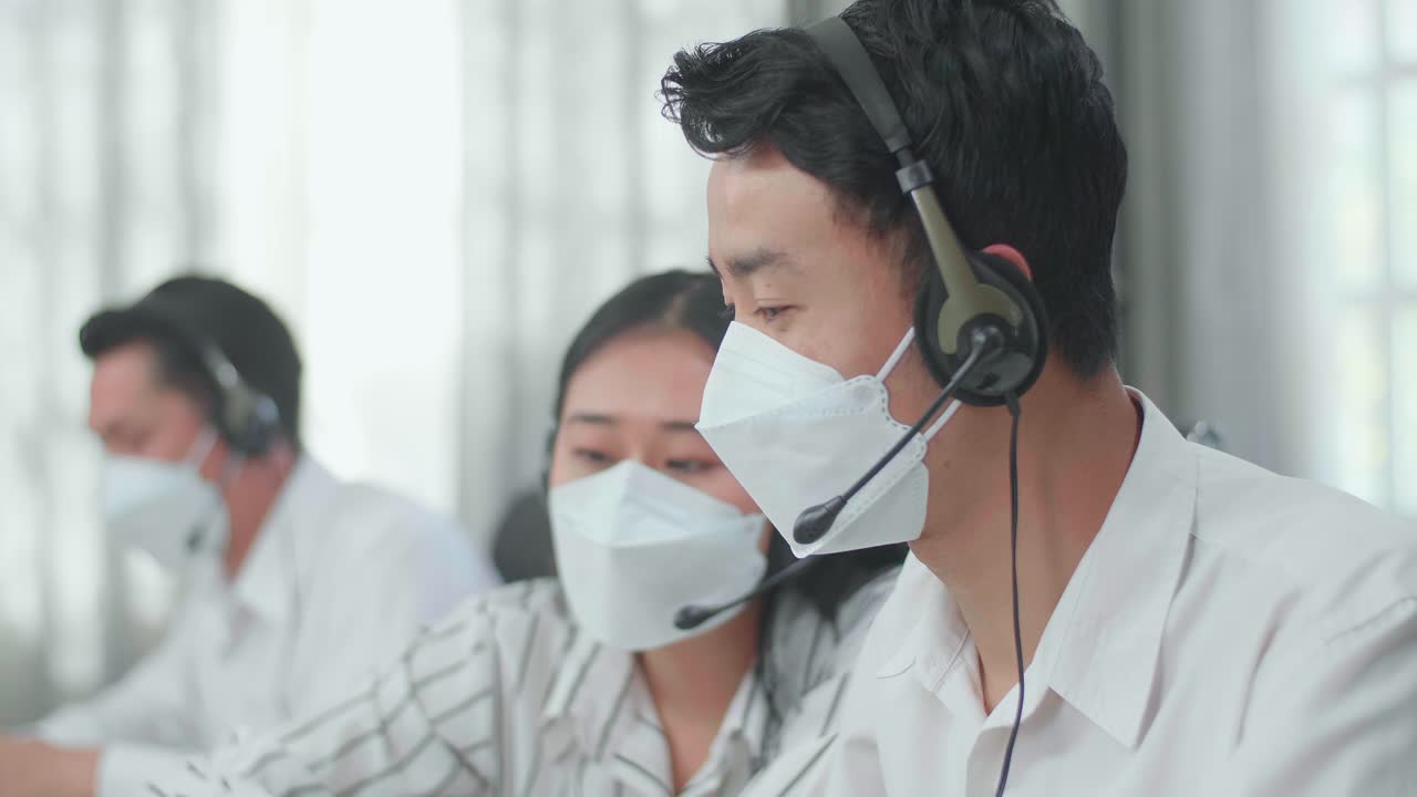 Close Up Of A Man And A Woman Of Three Asian Call Center Agents In Headsets And Masks Discussing About Work While Their Colleague Is Speaking With Customer On The Call At The Office