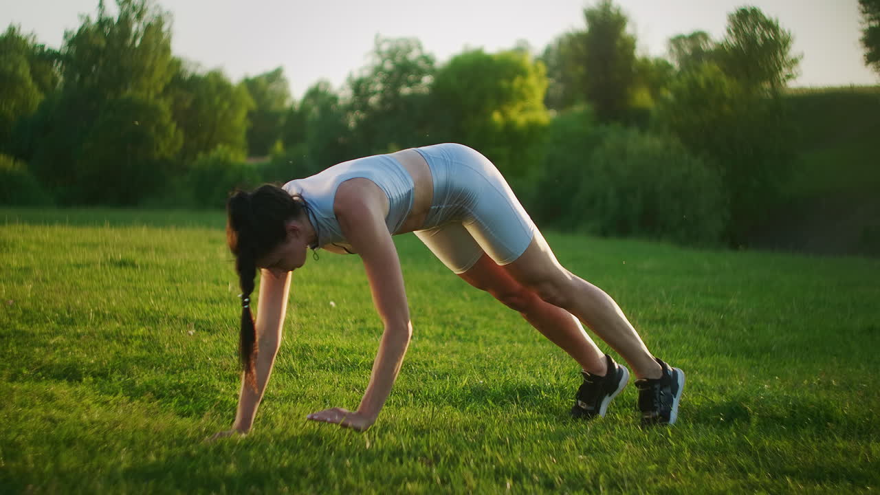 una mujer realiza un ejercicio de tabla de pie en la hierba al atardecer en un parque. lentamente va en las manos de en la hierba. levante las piernas en el ejercicio de tabla.