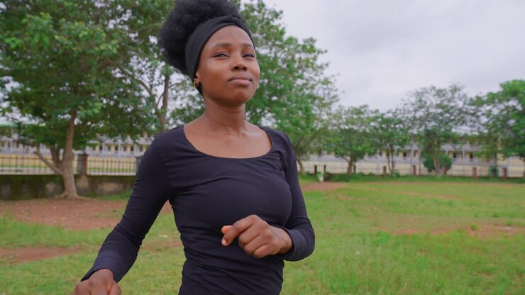 Una signora fa jogging in un parco con alberi verdi e un campo erboso aperto. La donna indossa un abbigliamento sportivo nero a maniche lunghe, inclusa una fascia per capelli, mentre fa jogging all'aperto, fitness all'aperto