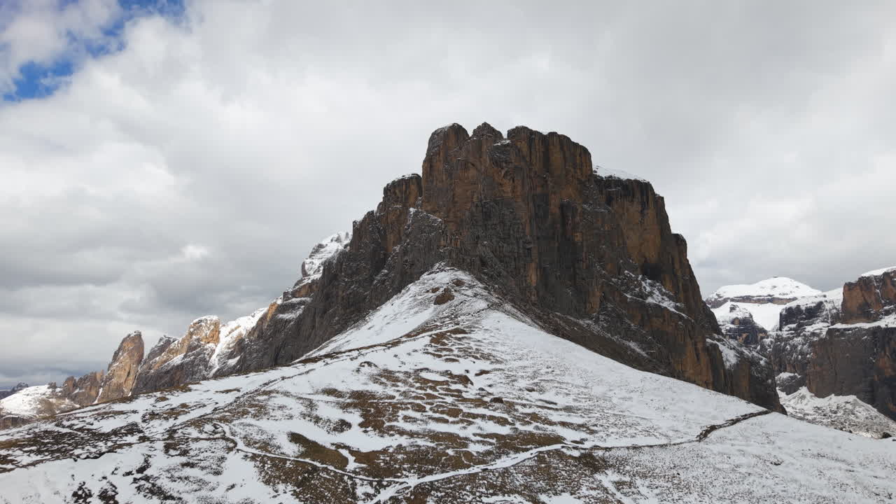 Aerial drone footage of a rugged rocky mountain partially covered in snow, with dramatic cliffs and cloudy skies in the Dolomites, Italy