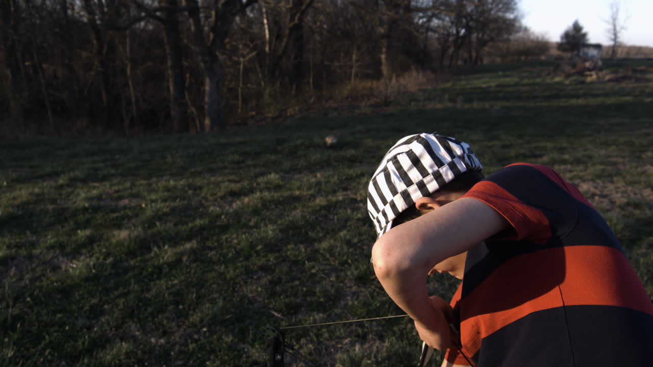 Young boy in striped cap practicing archery with bow in grassy field at sunset, Siloam Springs, Arkansas, side view