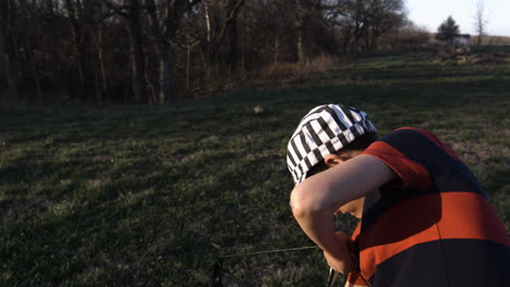 niño con gorra a rayas practicando tiro con arco en el campo cubierto de hierba al atardecer, siloam springs, arkansas, vista lateral