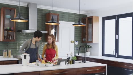 happy caucasian lesbian couple preparing food and using tablet in sunny kitchen