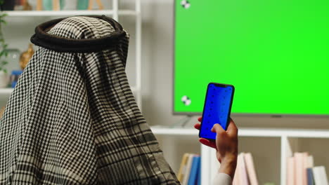 middle eastern man using phone with chroma key close-up. television with green screen in living room. wearing traditional islamic male clothes