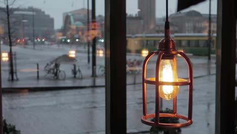 lantern lightbulb revealing behind leaves shadows, with rainy city in background and man passing with umbrella