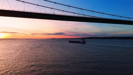 embark on a serene journey as a barge boat leisurely cruises beneath the humber bridge during sunset, enveloped in the golden hues of the setting sun, with the river awash in shimmering light