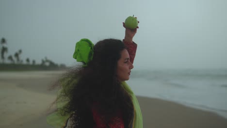 A fixed shot of a woman in green fabric holding a green apple above her head on the beach, her hair blowing in the wind