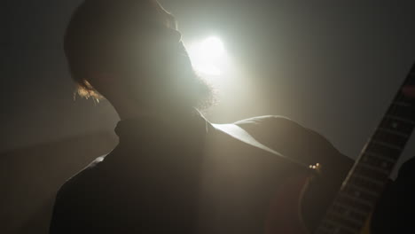 bearded guitarist in black, playing red guitar under spotlight in dark room