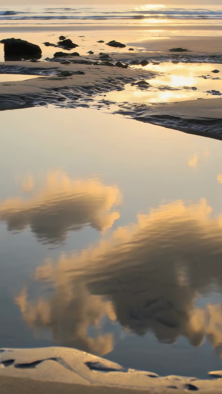 Vertical video: Cam pulling back, pool mirroring clouds and sun on beach, showing rills, rocks, sea