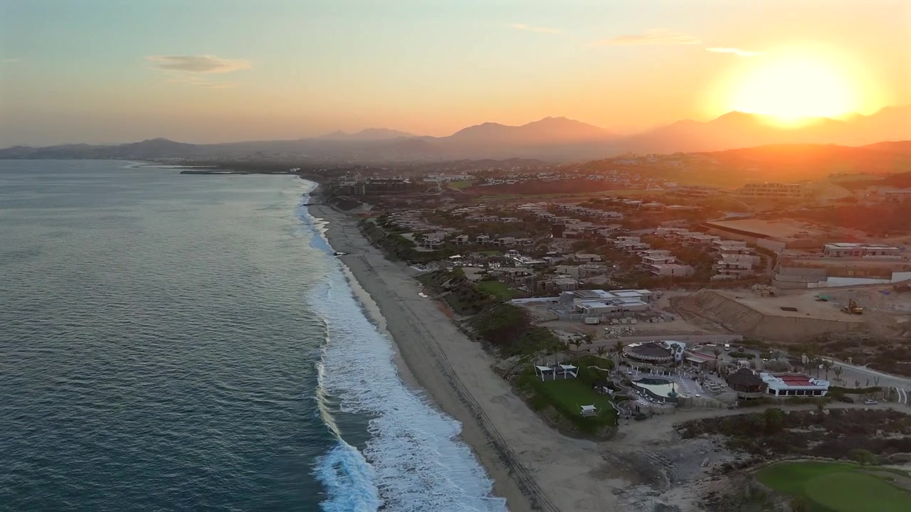 Drone recua ao entardecer sobre Puerto Los Cabos, com a comunidade residencial preenchendo o quadro abaixo e San Jose del Cabo e um pôr do sol vívido nas montanhas se estendendo pelo horizonte distante