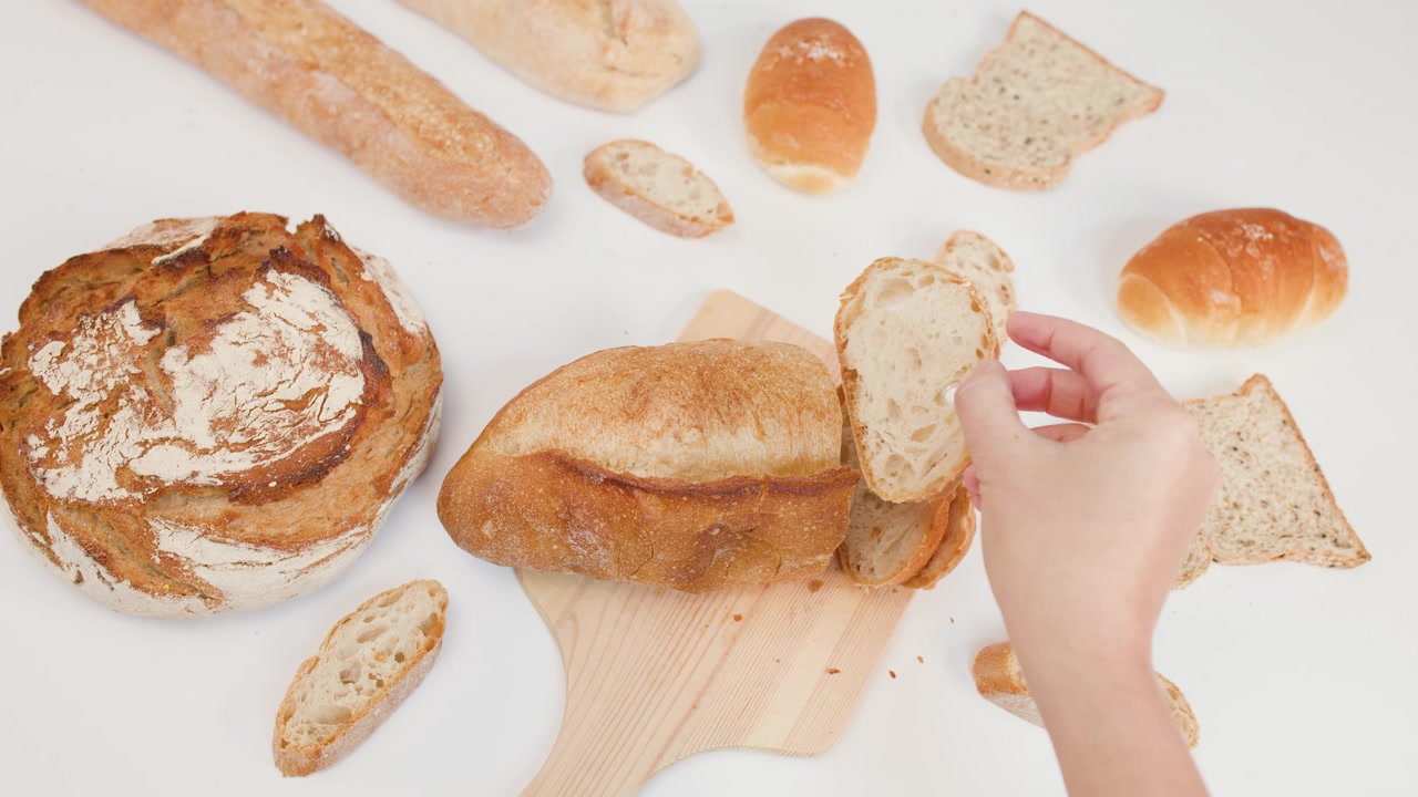 Overhead shot of hands slicing crusty sourdough bread on a wooden board against white background.