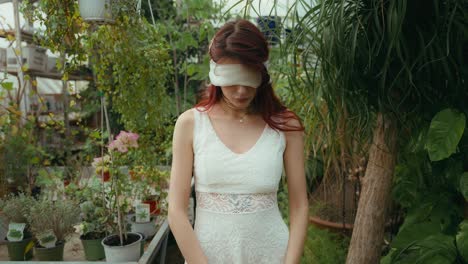 portrait dolly-in cinematic video of a young woman in white dress and blindfolded looking at camera in greenhouse of plants