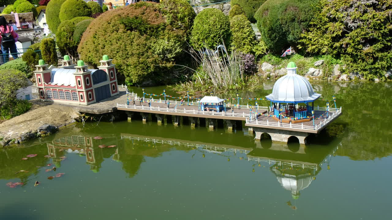 Wide Shot of Miniature Promenade Scene: Orchestra Performing in Bandstand Pavilion on Tranquil Green Lagoon with Reflections, Serene Waters, and Detailed Landscape in a Peaceful Miniature Setting
