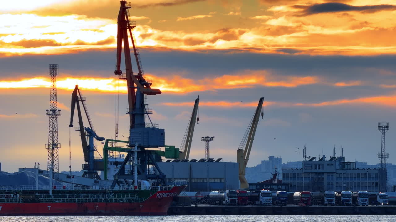 Sunset view of shipping yard with cranes and trucks in the harbor area. Trucks line up at the shipping yard as a ship docks at the harbor during sunset with cranes in view
