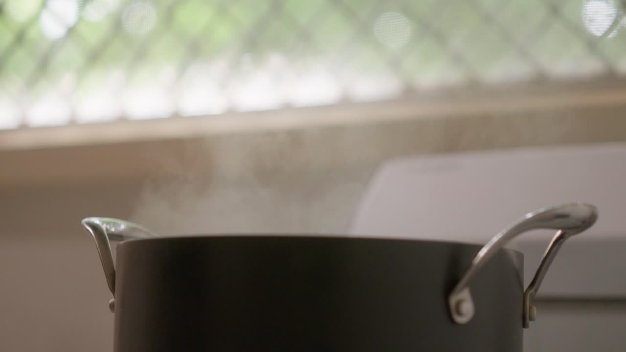 Simple, backlit shot of man's hand lifting the lid of a pot of boiling water sitting on a gas cooktop, and letting steam escape