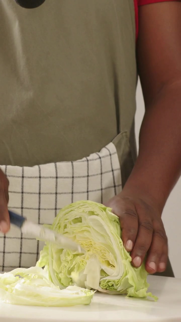 Vertical video: Holding halved cabbage, cook in apron shredding for salad at counter with knife