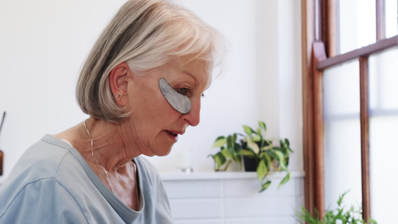 Senior woman applying eye patches at home, focusing on self-care routine