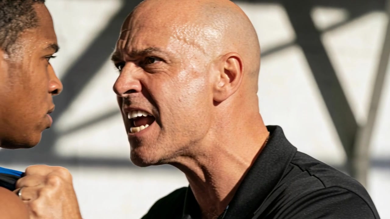 Advancing bald coach in black polo gripping athlete's blue singlet at gym, yelling over mistake