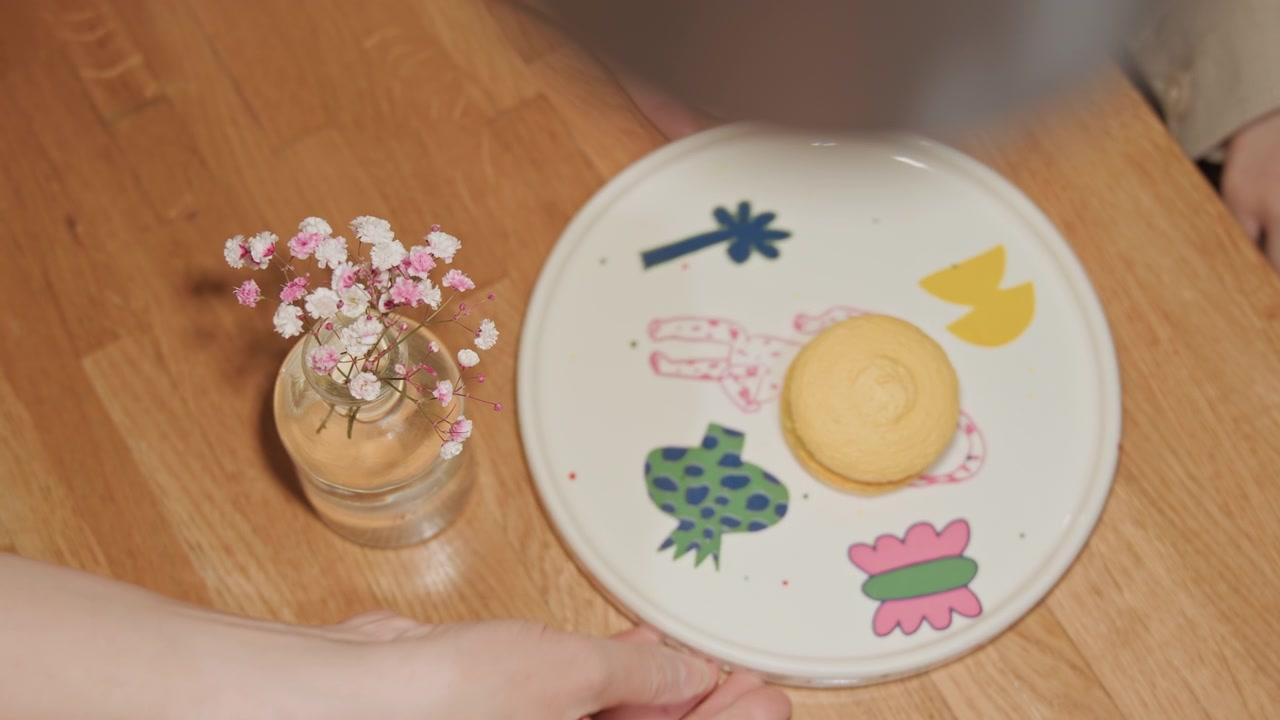 Close up of person hands reaching for and picking up decorative plate with colorful illustrations featuring pink flowers in glass vase on wooden table. scene includes soft lighting calm