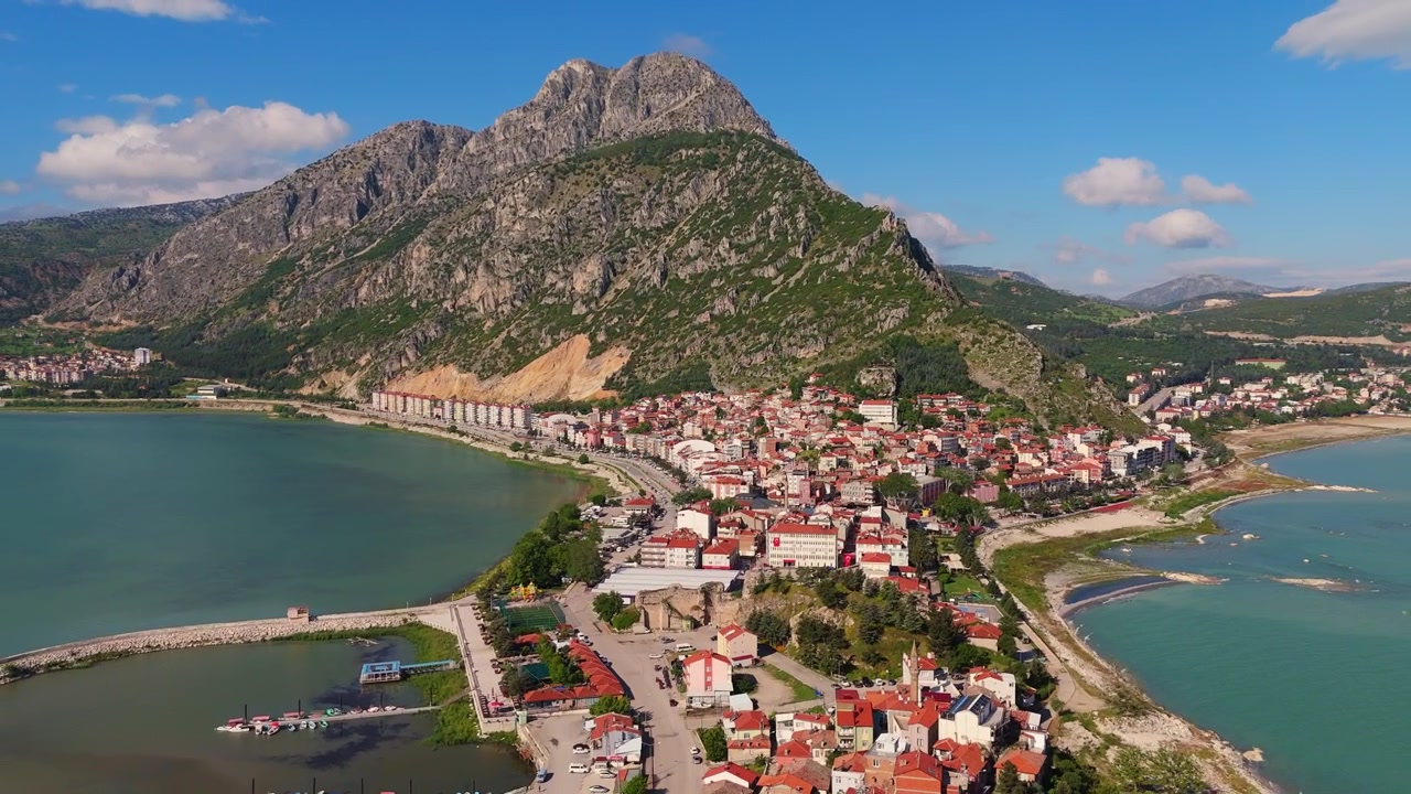 Eğirdir Gölü yarımada kasabasına tepeden bakan devasa kayalık dağ zirvesinin geniş panoramik drone çekimi. Türkiye'de görkemli jeolojik arazi ve turkuaz göl kenarı manzarası.
