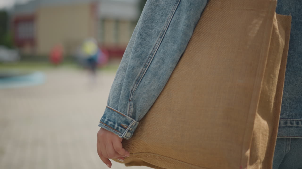 Casual woman carrying tote bag. Woman dressed casually carrying strawtextured bag outdoors in sunlight. Relaxed woman in denim jacket with woven straw bag strolling through suburban shopping plaza