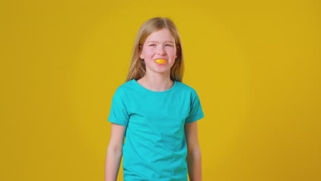 Retrato-De-Estudio-De-Una-Niña-Usando-Un-Segmento-De-Naranja-Para-La-Boca-Y-Los-Dientes-Contra-Un-Fondo-Amarillo