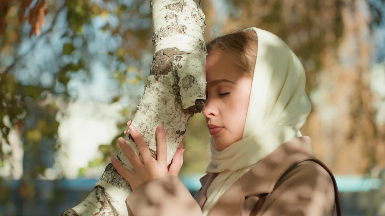Donna caucasica che tocca il tronco di una betulla, luce solare autunnale, foulard color crema, cappotto beige, occhi chiusi, bacio delicato sulla corteccia, ricordo emotivo, bokeh morbido, ambientazione tranquilla del parco, toni caldi cinematografici.