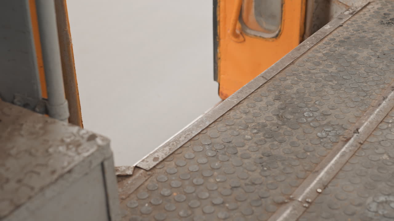 Primo piano dello spazio tra la porta arancione del treno e la piattaforma che mostra un gradino di metallo usurato e piastre tattili sul pavimento accanto alla porta arancione della metropolitana, vernice scrostata e bordi arrugginiti, texture industriale granulosa, atmosfera urbana ambientale