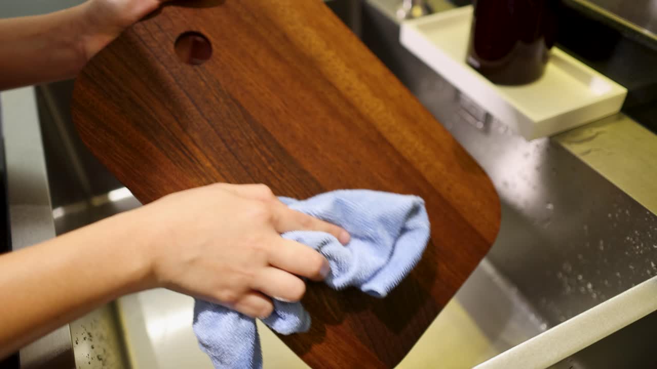 Primo piano di mani che puliscono un tagliere di legno sopra un lavello da cucina con un panno in microfibra.