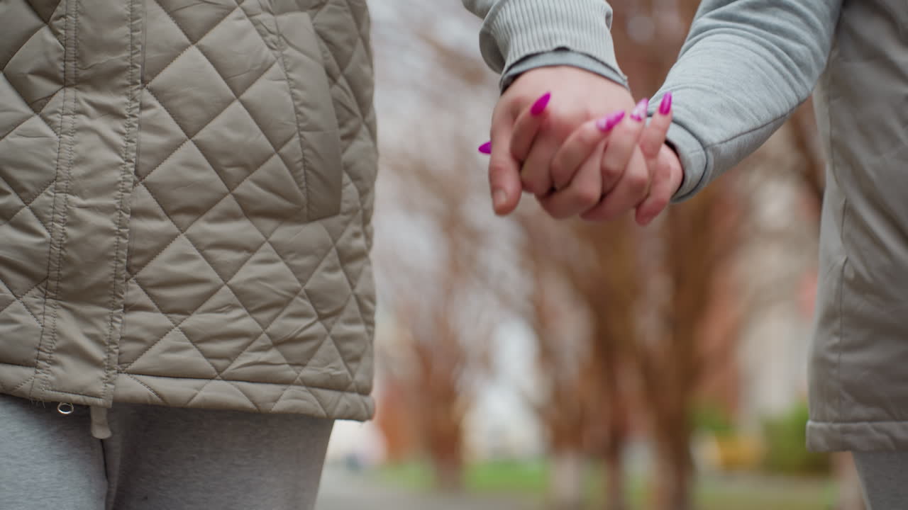 Close-up of friends walking with hands locked together, gently swinging while strolling, wearing cozy outfits with quilted vest and hoodie, blurred trees and soft colors in background