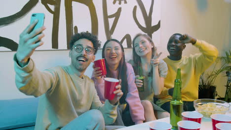 funny multiethnic young friends taking a selfie video while sitting on sofa and having a beer party at home