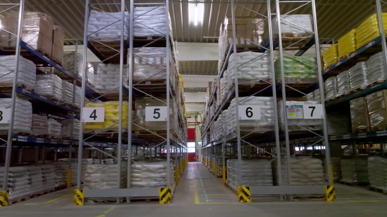 Toma panorámica dentro de un almacén industrial con estanterías de paletización altas, mercancías embaladas, pasillos de almacenamiento e infraestructura logística para temas de distribución, cadena de suministro e inventario.