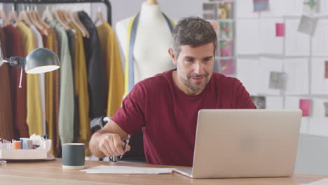 mature male fashion designer in wheelchair in studio working on laptop computer and drinking coffee