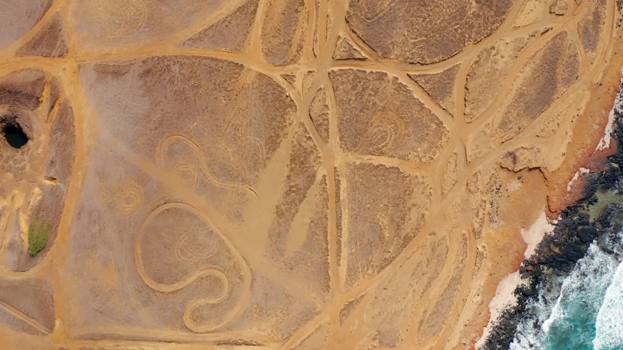 A vertical aerial view of looping tire tracks on an arid coastal plain beside rocky shoreline and foaming waves