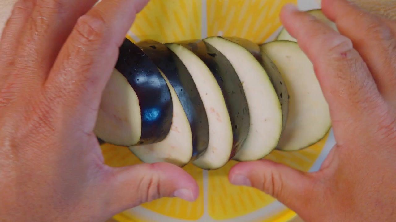 Vue plongeante de mains masculines coupant une aubergine violette fraîche avec un couteau aiguisé sur une planche à découper jaune, préparant les ingrédients pour un repas végétarien sain dans la cuisine à la maison