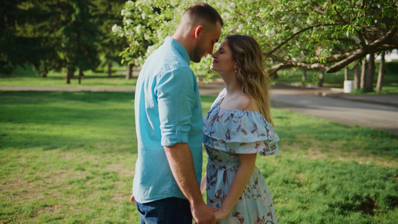 Paar hält Händchen in sonnigem Park, lächelt, teilt einen zärtlichen Moment. Bäume blühen, Gras üppig, Kind spielt im Hintergrund. Ideal für romantische Szenen, Hochzeiten oder Familienveranstaltungen.