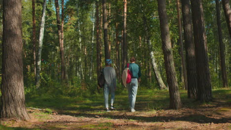 two women walking through lush forest, one with blue hair tie and backpack, other with cloth draped on bag, hair tied back, surrounded by tall trees and dappled sunlight